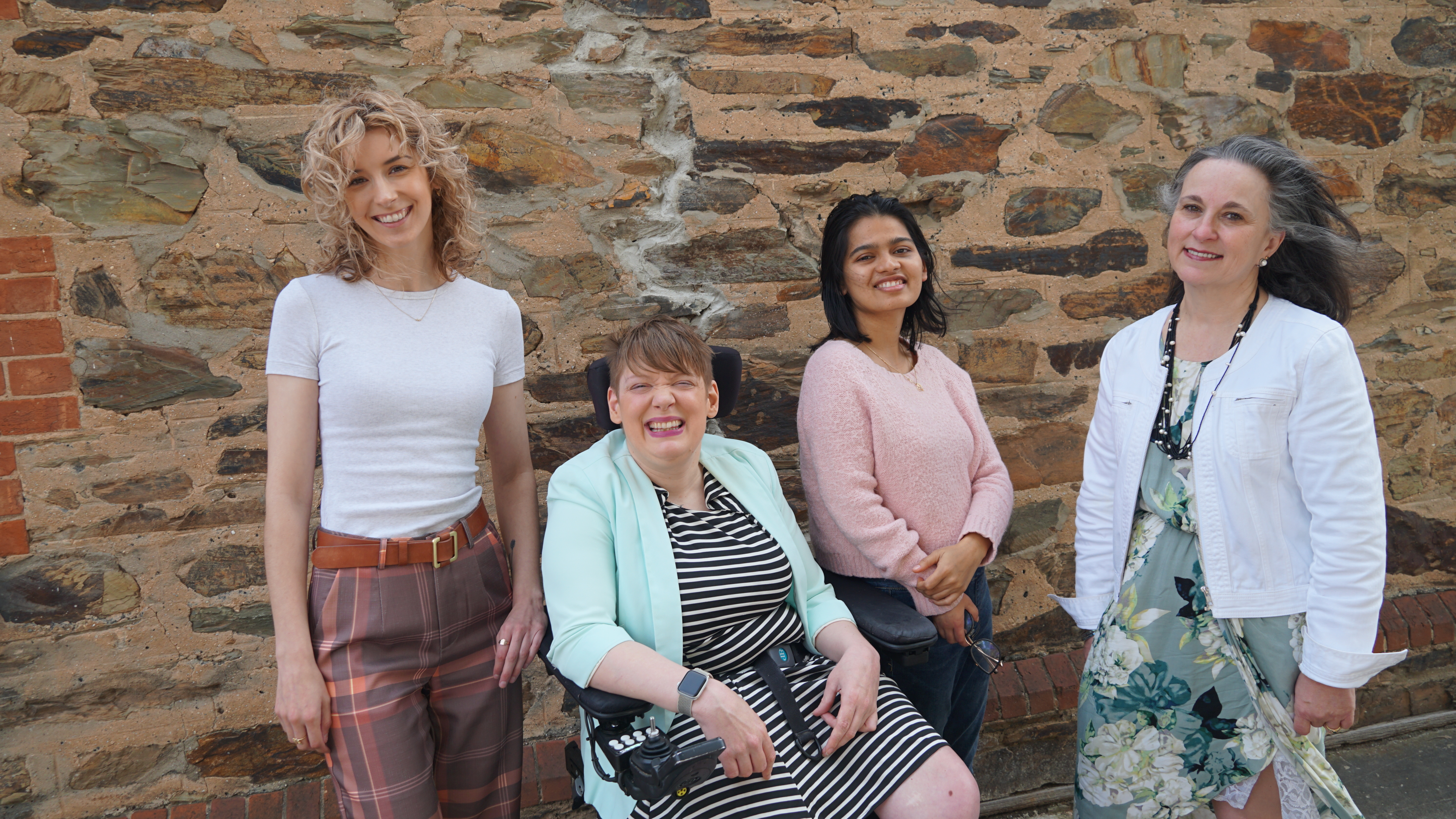 Four people posing in front of a brick wall. A white woman with blonde curly hair wearing a white t-shirt and plaid pants, standing next to a white person in a wheelchair with a brown pixie haircut, wearing a striped dress and aqua blue blazer. Another wo